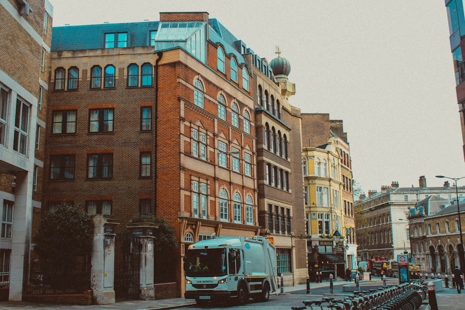 A street scene showing a white waste collection truck with visible rust and mechanical components at the rear, positioned on a cobblestoned pavement alongside old multi-story buildings with weathered